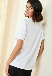 Haut blanc à manches courtes avec une coupe décontractée. Tissu doux et lisse ; encolure ronde ; sans motifs visibles. Le modèle se tient devant un fond clair.