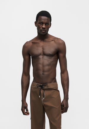 Mężczyzna o umięśnionej sylwetce z krótkimi włosami, ubrany w brązowe szorty ze sznurkiem, stoi na jasnym, jednolitym tle z neutralnym wyrazem twarzy.