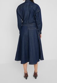 Dunkelblaues Denim-Hemdkleid mit langen Ärmeln, tailliertem Schnitt, ausgestelltem Rock und Details an der Gesäßtasche. Saumlinie leicht abgerundet.