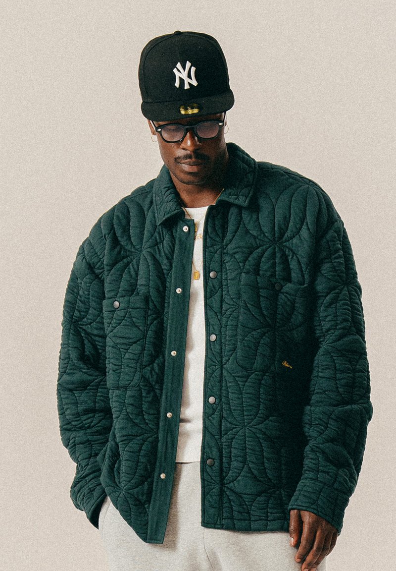 Άνδρας που φοράει μαύρο καπέλο New York Yankees, στρογγυλά γυαλιά, πράσινο καπιτονέ μπουφάν, λευκό πουκάμισο και γκρι παντελόνι μπροστά σε απλό μπεζ φόντο.