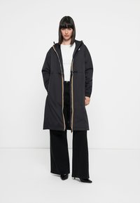 Young woman wearing a long black hooded coat over white top and black wide-leg pants, standing against a white background.