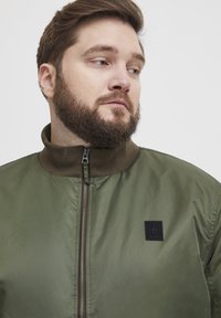 Maslinasto zelena bomber jaka s visokim ovratnikom, zatvaranjem na cipzaru i teksturiranom tkaninom. Ima crnu markicu s logotipom s lijeve strane.