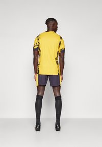 Gele sporttrui met zwarte accenten en geometrische patronen, gecombineerd met donkere shorts en zwarte sokken, ontworpen voor atletische prestaties.