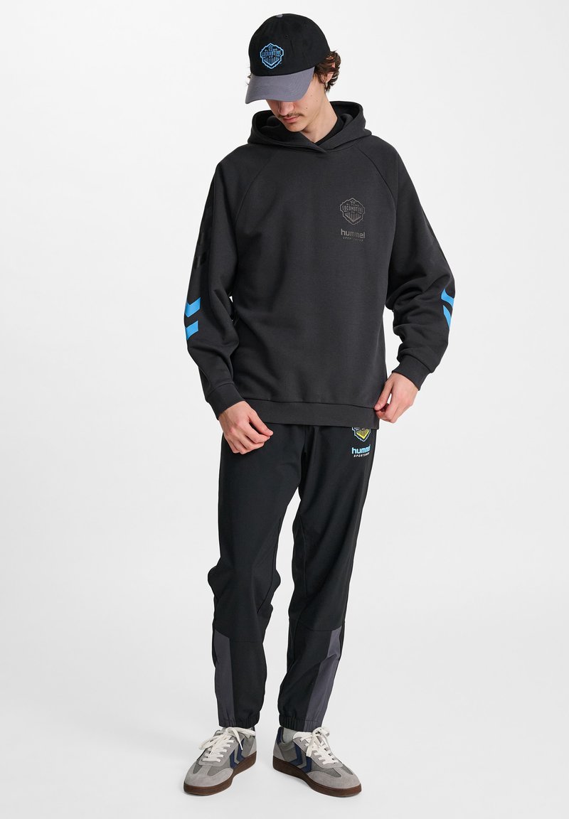 Svart hoodie med blå accenter och logotyp, tillsammans med svarta byxor med blå och grå paneler. Vita sneakers fulländar outfiten.