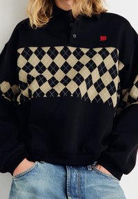 Pull noir avec un col à revers, présentant un motif losange beige sur la poitrine et un détail de logo rouge. Tissu doux avec ourlet côtelé.