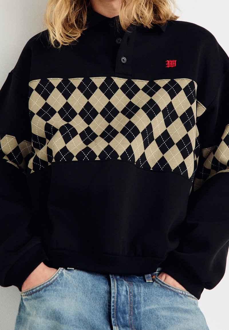Pull noir avec un col à revers, présentant un motif losange beige sur la poitrine et un détail de logo rouge. Tissu doux avec ourlet côtelé.