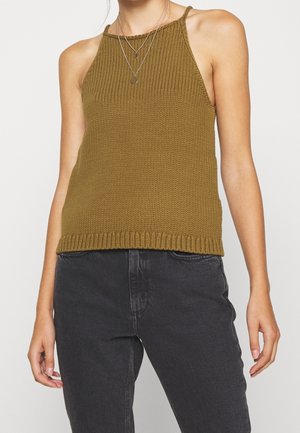 Person wearing olive green knitted sleeveless top and black high-waisted jeans, accessorized with layered silver necklaces.