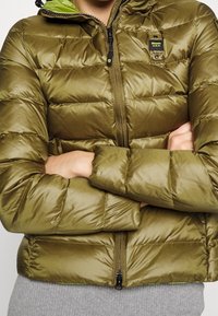 Persona con chaqueta acolchada verde oliva con cremallera frontal y brazos cruzados, mostrando el parche con el logo de Blauer U.S.A. en el bolsillo del pecho.