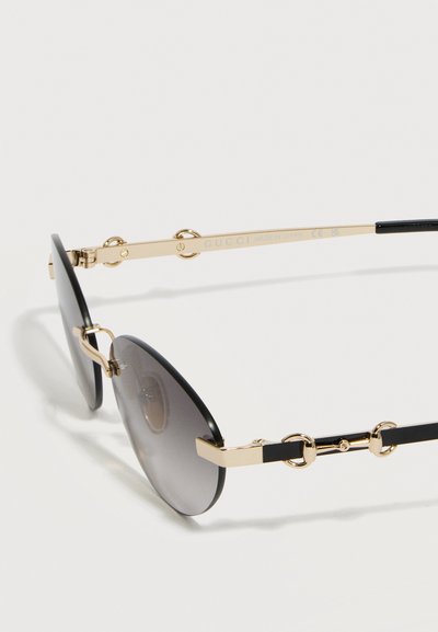 Μαύρα στρογγυλά γυαλιά ηλίου με χρυσούς μεταλλικούς βραχίονες που διαθέτουν διακοσμητικά στοιχεία με διαπλεκόμενους κρίκους και φακούς με σκίαση βαθμιαίας μετάβασης σε σκούρο χρώμα σε λευκό φόντο.