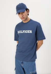 Nuori mies, jolla on päässään sininen hattu, sininen "Hilfiger"-t-paita ja siniset farkut, seisoo yksinkertaista valkoista taustaa vasten.