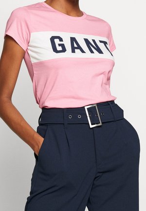 Frau trägt ein pinkes GANT-T-Shirt, das in hoch taillierte marineblaue Hosen mit einem breiten Gürtel und eckiger silberner Schnalle gesteckt ist, Hände in den Taschen.