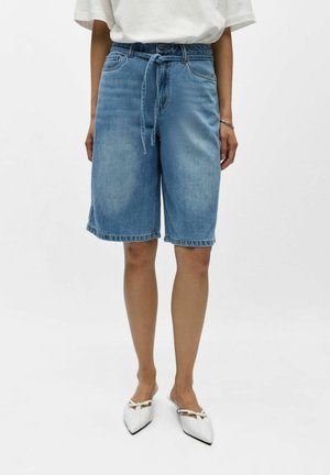 Persona con pantalones cortos de mezclilla azul claro hasta la rodilla, con un cinturón de lazo en la cintura, zapatos blancos puntiagudos y una camiseta blanca de manga corta.