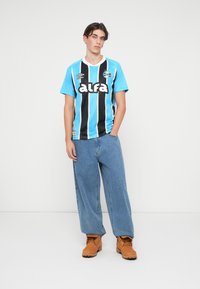 Camiseta de fútbol a rayas azul y negra con el logo "alfa", combinada con pantalones de mezclilla de pierna ancha en azul claro y botas marrones con cordones.