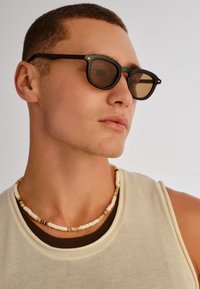 Νεαρός άνδρας που φοράει μαύρα γυαλιά ηλίου, περιδέραιο με χάντρες και μπεζ αμάνικο τοπ, κοιτάζοντας προς τα πλάγια μπροστά σε απλό φόντο.