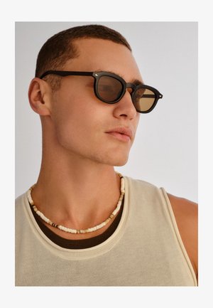 Noor mees, kes kannab musta päikeseprille, helmestega kaelakeed ja beežikat lühikeste varrukatega topi, vaatab küljele ilma taustata.