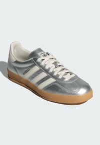 Silberner Sportschuh mit glattem Obermaterial, weißen Lederstreifen und einer Gummisohle. Ausgestattet mit einem Schnürdesign und dezentem Branding auf der Zunge.