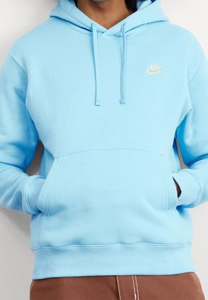 Person trägt einen hellblauen Nike-Hoodie mit Fronttasche und Kordelzug, Hände in der Tasche, kombiniert mit braunen Hosen.