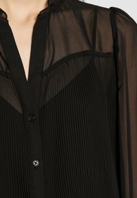 Musta läpinäkyvä blouse, jossa on pystysuorat laskokset, nappikiinnitys ja mandariinikaulus. Kankaassa on pehmeä, kevyt rakenne.