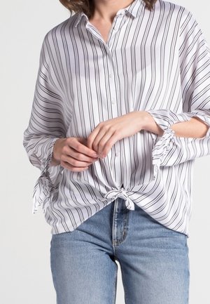 Vrouw draagt een witte blouse met zwarte verticale strepen, gestrikt ter hoogte van de taille, en lichtblauwe jeans, handen losjes gevouwen voor zich.