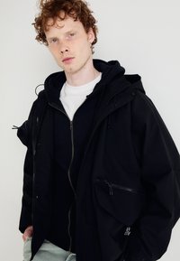 Black outer jacket with a hood, zippered pockets, layered over a black zip hoodie and white t-shirt. Textured material and relaxed fit.