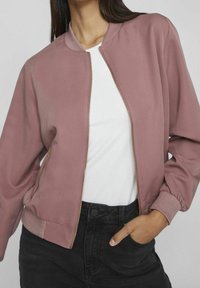 Woman wearing a mauve bomber jacket over a white shirt with one hand in the pocket of her high-waisted black jeans against a plain background.
