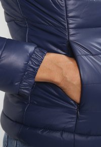 Granatowa kurtka puchowa o pikowanej fakturze, z elastycznymi mankietami i bocznymi kieszeniami. Materiał wydaje się gładki i błyszczący, o dopasowanej sylwetce.