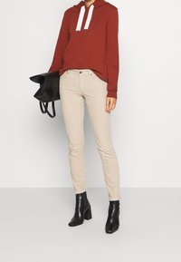 Felpa rossa con cappuccio e cordini bianchi, pantaloni beige aderenti e stivaletti neri con tacco, tiene in mano una borsa nera con bordi arrotolati.