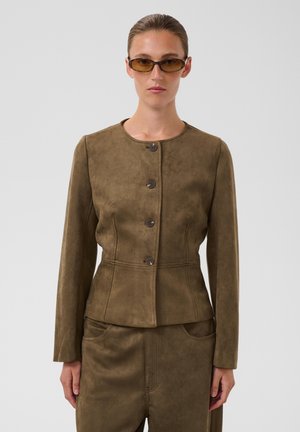 Femme portant une veste en suède marron boutonnée et un pantalon assorti coupe slim avec de petites lunettes de soleil marron sur fond uni.