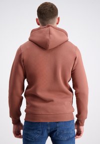 Brūna kapuce ar teksturētu apdari un ribotām piedurknēm, kopā ar ziliem džinsiem ar aizmugurējo kabatu izšuvuma detaļām.