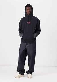 Un sweat à capuche noir avec un logo rose, associé à un jean en denim noir ample et des baskets crème, avec une coupe décontractée et des poignets.