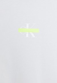 Světlé šedé tričko s bílým nápisem "cK" a jasně žlutým vodorovným pruhem přes logo. Vyrobeno z měkké látky s hladkou texturou.