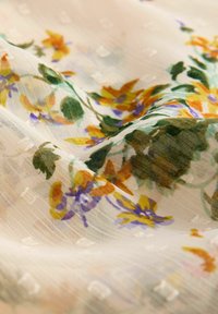 Tissu beige transparent avec des points blancs brodés et un motif floral coloré dans des tons de vert, jaune, violet et orange.