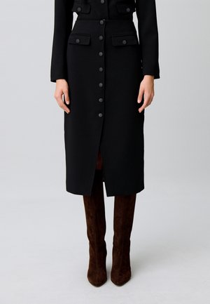 Persona che indossa una gonna midi nera con bottoni e tasche e stivali in suede marrone scuro fino al ginocchio, in piedi su uno sfondo semplice.