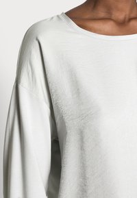 Light gray long-sleeve top with a loose fit, featuring a wide neckline and textured fabric. Seam detailing at the shoulders.