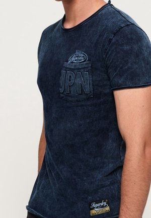 Homme portant un t-shirt bleu foncé délavé avec une poche poitrine portant la mention "JPN" et un petit logo "Superdry" près de l'ourlet.