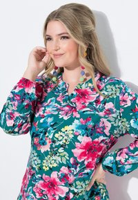Mujer con cabello rubio largo que lleva una blusa floral colorida y pendientes de colores a juego, sonriendo y posando con la mano cerca de la cara.