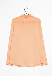 Una camicia a maniche lunghe di colore pesca, realizzata in un tessuto morbido e leggero, con colletto classico e polsini con bottoni, appesa a un gancio nero.