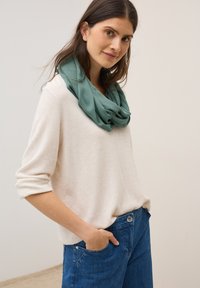 Pull en tricot beige clair à manches roulées, associé à un jean en denim bleu. Une écharpe verte drapée et surdimensionnée repose autour du cou.