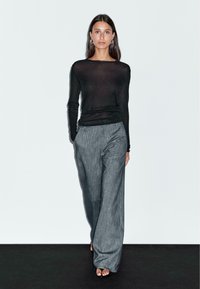 Black sheer long-sleeve top paired with loose, light gray, textured wide-leg pants. Model stands against a plain background, barefoot.