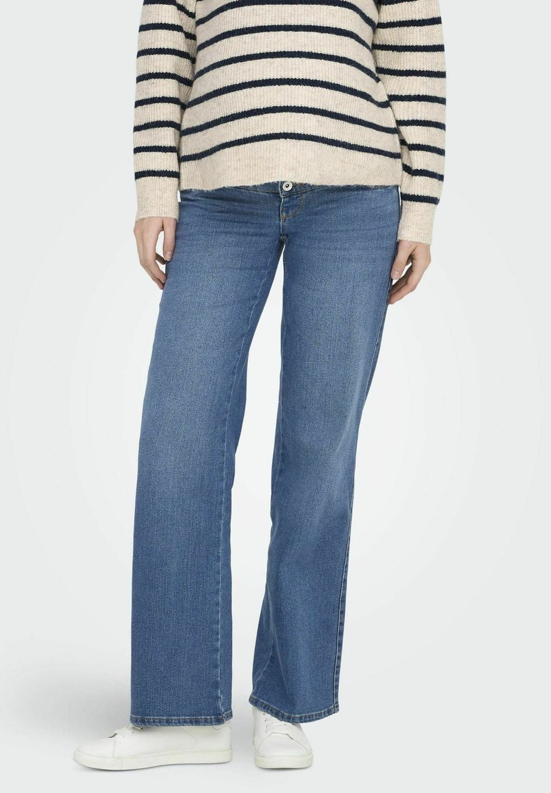 Blaue weit geschnittene Jeans mit einem klassischen Fünf-Taschen-Design und einem Knopfverschluss, kombiniert mit einem gestreiften Pullover in Creme und Marineblau sowie weißen Schuhen.