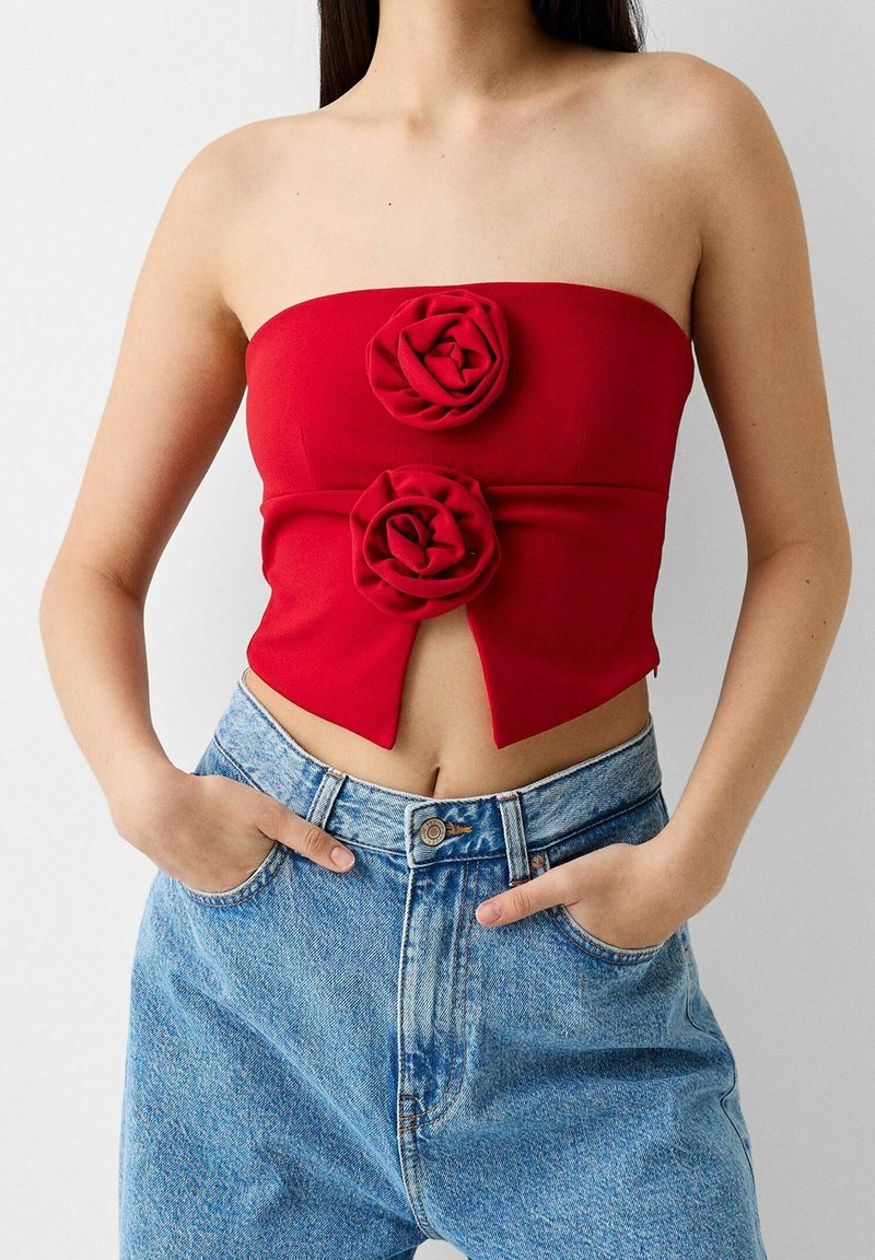Rotes trägerloses Crop Top mit zwei strukturierten Rosenapplikationen auf der Vorderseite; kombiniert mit hochtaillierten hellblauen Jeans.