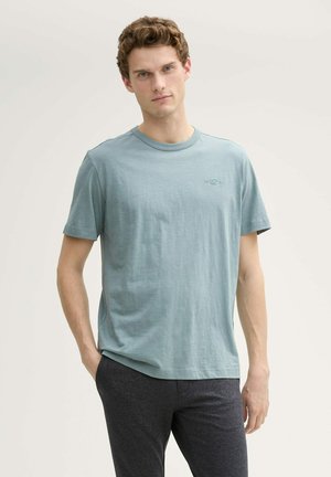 Helles blaues T-Shirt mit kurzen Ärmeln, Rundhalsausschnitt, aus weichem Baumwollmaterial, mit einem kleinen Logo auf der Brust, kombiniert mit dunkelgrauen Hosen.
