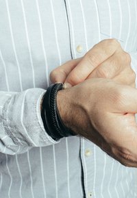 Fekete bőrből készült, fonott mintázatú karkötő, ami a csuklón viselve, világoskék csíkos ing háttér előtt látható, kiemelve annak kialakítását és kontrasztját.
