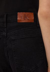 Detailný záber na zadnú vrecko čiernych džínsoch z denimu s hnedou koženou nášivkou loga Calvin Klein a časťou ruky osoby v čiernom tričku.