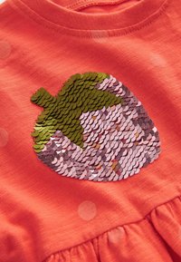 Motif de gland en sequins verts et roses sur tissu corail avec de faibles pois blancs et couture froncée en bas.