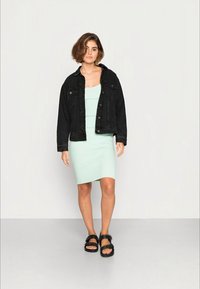 Jaqueta de ganga preta sobre um vestido canelado em verde-menta claro, que chega até à metade da coxa. Sandálias pretas com tiras. Fundo simples.