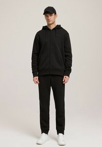 Czarny hoodie na zamek z troczkami, kieszeniami z przodu i pasującymi czarnymi dresami, uzupełniony białymi sneakersami i czapką w kolorze czarnym.