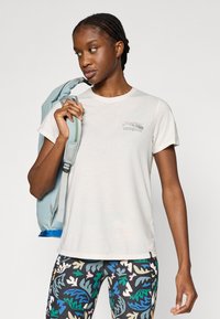 Ανοιχτόχρωμο μπλουζάκι Patagonia με κοντά μανίκια και γραφικό βουνό· συνδυασμένο με μαύρο κολάν με πολύχρωμα σχέδια φύλλων· κρατάει μπλε σακίδιο πλάτης.