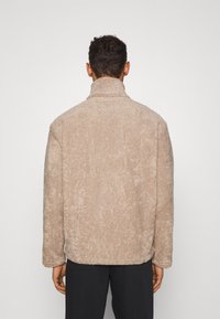 Lysbrun fleecejakke med høj krave, blød tekstur og afslappet pasform. Har et enkelt design og minimal dekoration på ryggen.