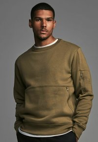 Sudadera verde oliva, con cuello redondo, puños con ribete, bolsillo lateral con cremallera y tejido texturizado con un acabado suave.